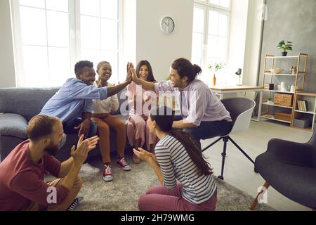 Due uomini interrazziali si danno cinque a vicenda mentre si siedono in compagnia di amici a casa. Foto Stock