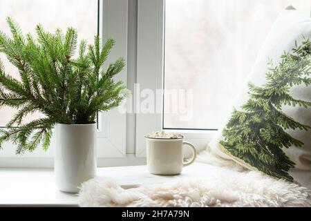 Rami di abete naturale come albero di Natale in vaso e tazza di caffè su inverno accogliente davanzale con cuscino e pelle di pelliccia. Natale romantico moody. Foto Stock