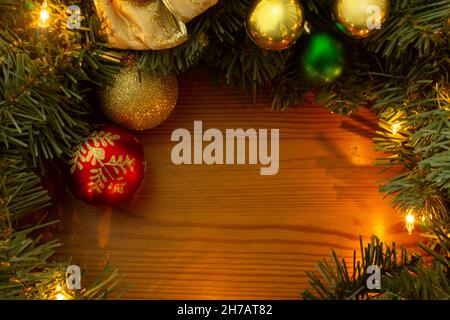 Cornice formata da rami di albero di Natale verde, luci gialle, lampadine dorate e rosse su un tavolo di legno Foto Stock