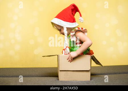 La bambina in cappello di Babbo Natale e un costume assistente di elfo siede in cartone scatola. Concetto di ricezione o spedizione pacco di Natale a casa. Consegna ser Foto Stock