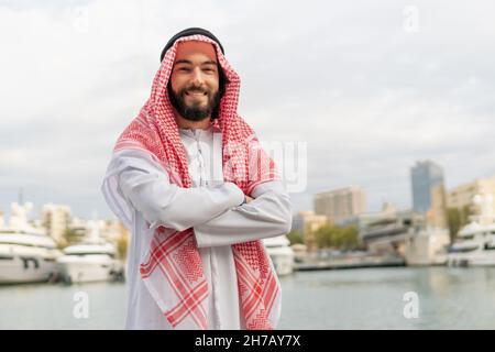 Ritratto di felice uomo arabo allegro in abiti tradizionali keffiyeh e bianco thibe sorridente a macchina fotografica mentre si trova in porto, tenendo le braccia incrociate. Foto del riuscito uomo d'affari saudita Foto Stock