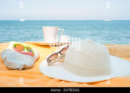 cappelli su un asciugamano sulla spiaggia. cappelli per bambini e donne su un asciugamano arancione con una tazza su sfondo marino Foto Stock