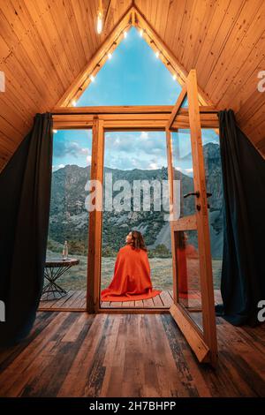 Donna sola sul portico di una piccola casa di legno in montagna. Il concetto di glamping e vacanze idilliache Foto Stock