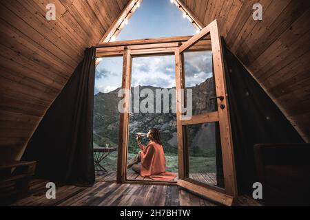 Donna sola bevendo acqua sul portico di una piccola casa di legno in montagna. Il concetto di glamping e vacanze idilliache Foto Stock