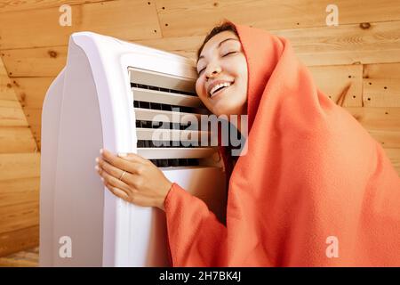 Donna divertente si riscalda ad un radiatore elettrico nella sua casa di legno in inverno, avvolto in una coperta. Il concetto di energia e comfort nella stagione fredda Foto Stock