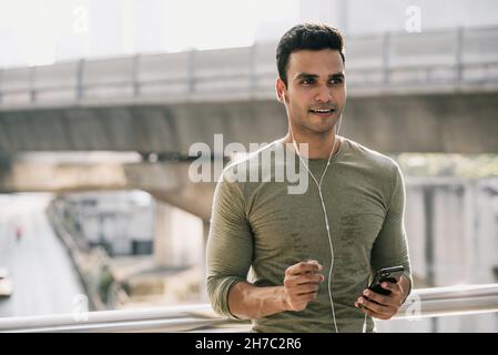 Sorridente giovane sudato bell'uomo indiano che ascolta musica su smartphone all'aperto in città dopo il jogging Foto Stock