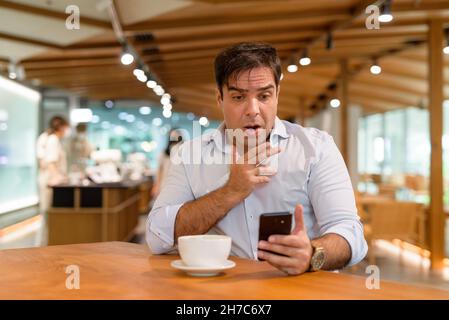 Scioccato e sorpreso uomo persiano seduto al bar mentre si utilizza il telefono cellulare orizzontale scatto Foto Stock