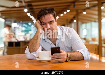 Ritratto di un bel uomo persiano seduto al bar mentre si usa il telefono cellulare Foto Stock