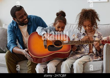 Ritratto di felice padre afroamericano che suona la chitarra con due bambine a casa Foto Stock