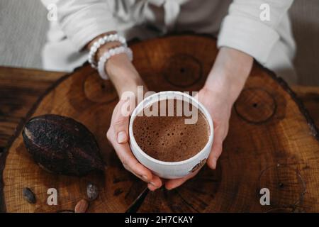 Cacao cerimoniale caldo fatto a mano in tazza bianca. Donna mani che tengono cacao artigianale, vista dall'alto su tavola di legno. Bevanda biologica sana al cioccolato preparata da Foto Stock