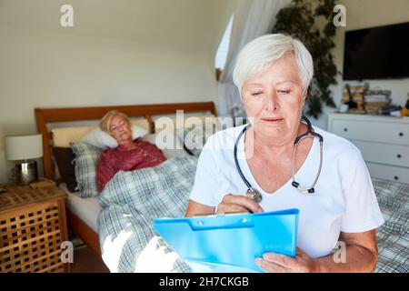 Il medico femminile guarda i file dei pazienti in casa di cura durante la visita a casa con anziani in background Foto Stock
