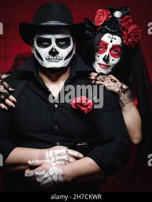 bella donna con un trucco del cranio dello zucchero con una corona di fiori sulla sua testa e un uomo dello scheletro in un cappello nero. Coppia su sfondo rosso scuro Foto Stock