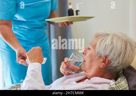 La donna anziana prende una tavoletta con un sorso d'acqua, sotto la supervisione del personale infermieristico a casa Foto Stock