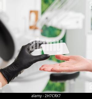 Primo piano della mano di stomatologo femminile in guanti sterili neri che danno il biglietto da visita della clinica al paziente. Dottore che consegna il biglietto di visita della giovane donna. Concetto di odontoiatria, cura dentale e stomatologia. Foto Stock