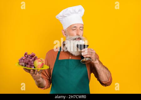 Ritratto di bearded handsome sognante uomo chef grigio-capelli bere vino mangiare snack isolato su sfondo di colore giallo vivo Foto Stock