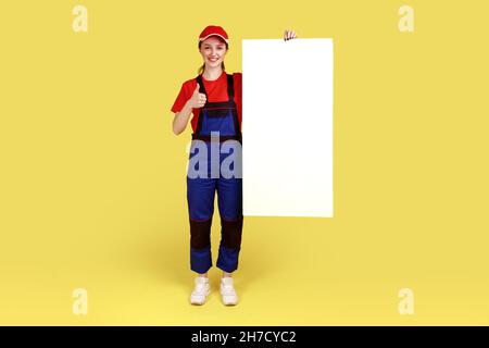 Ritratto a tutta lunghezza di donna lavoratore in piedi con poster vuoto con spazio copia per la vostra pubblicità, mostrando pollice su, indossare tute e cappuccio rosso. Studio interno girato isolato su sfondo giallo Foto Stock