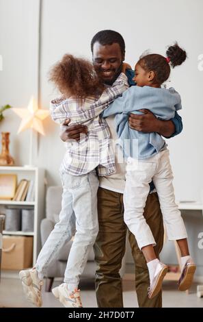Ritratto verticale di felice padre afroamericano che tiene due simpatiche figlie a casa Foto Stock