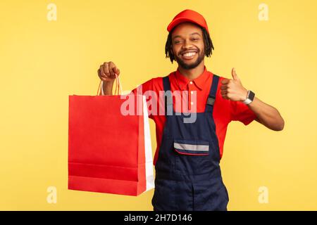 Haappy corriere attraente indossando tute blu e T-shirt rossa che tiene le borse per lo shopping e mostrando i pollici in su, consiglia servizio di consegna rapida. Studio interno girato isolato su sfondo giallo. Foto Stock