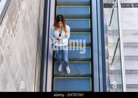 Ragazza allegra con smartphone che cammina sulla scala mobile mentre sorride, foto dall'alto. Concetto di studente Foto Stock