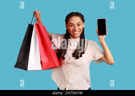 Donna felice positiva con dreadlocks che tiene i sacchetti di shopping nelle mani e che mostra il telefono delle cellule con l'esposizione vuota per l'annuncio, indossando la camicia bianca. Studio interno girato isolato su sfondo blu. Foto Stock