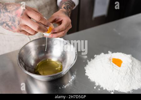 Vista croppata dello chef che tiene l'uovo vicino al recipiente e la farina sul tavolo da cucina Foto Stock