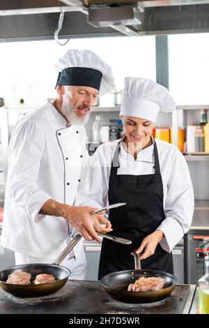 Chef che tiene le pinze vicino a un collega sorridente e carne sulle padelle in cucina Foto Stock