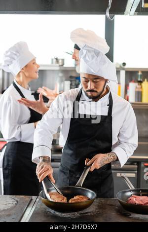 Lo chef asiatico prepara la carne sul piano cottura mentre i colleghi offuscati parlano in cucina Foto Stock