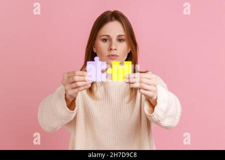 Ritratto di seria concentrata attraente bionda donna che tiene giallo e viola pezzi di puzzle, risolvere compiti, indossare maglione bianco. Studio interno girato isolato su sfondo rosa. Foto Stock