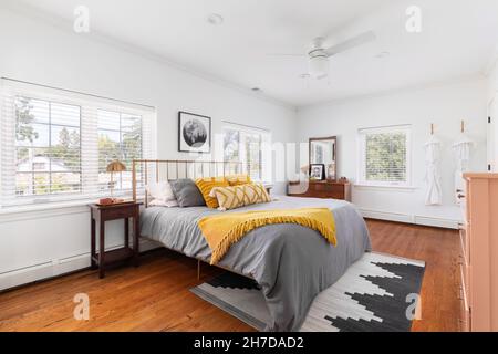 Una luminosa camera da letto con pareti bianche, biancheria da letto gialla e grigia e un tappeto su pavimenti in legno. Foto Stock