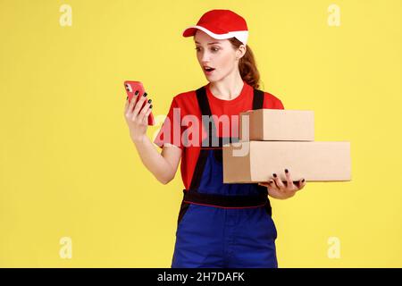 Ritratto di eccitata donna di consegna stupita tenendo i pacchi in mani, utilizzando il telefono per controllare l'indirizzo, avendo errore con la consegna. Studio interno girato isolato su sfondo giallo. Foto Stock