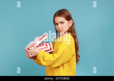 La vista laterale della bambina apre la scatola del regalo, esprime tristezza, celebra la vacanza, deluso di regalo, indossando il maglione giallo casual stile. Studio interno girato isolato su sfondo blu. Foto Stock