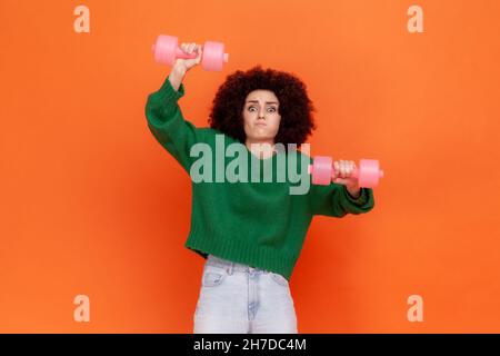 Donna sportiva con acconciatura afro indossando un maglione verde casual facendo esercizi sportivi per le braccia, utilizzando manubri duri, divertente espressione facciale. Studio interno girato isolato su sfondo arancione. Foto Stock