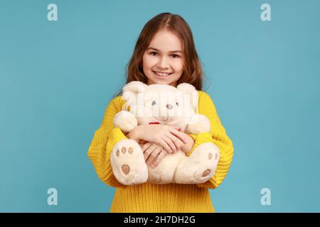 Ritratto di bambina che abbraccia l'orso giocattolo bianco carino, guardando la macchina fotografica, sognando, godendo presente, indossando il maglione giallo casual stile. Studio interno girato isolato su sfondo blu. Foto Stock