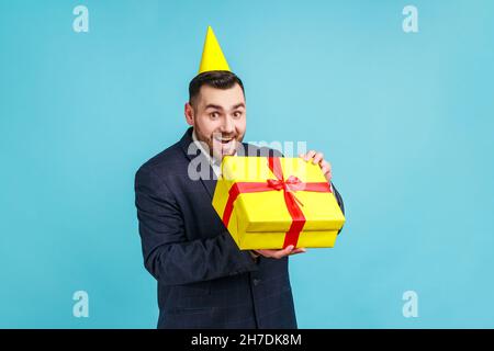 Un uomo d'affari eccitato che indossa un abito ufficiale e un cono da festa estremamente felice con il regalo di vacanza, i sogni si avverano, guardando la fotocamera. Studio interno girato isolato su sfondo blu. Foto Stock