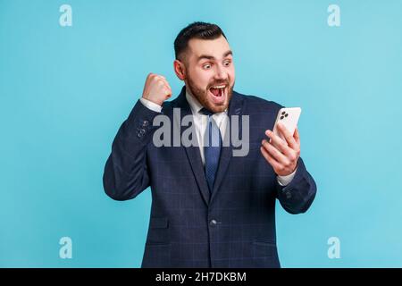 Uomo felice soddisfatto con barba in abito stile ufficiale con smartphone e sorridente facendo sì gesto, celebrando la lotteria online o la vittoria omaggio. Studio interno girato isolato su sfondo blu. Foto Stock