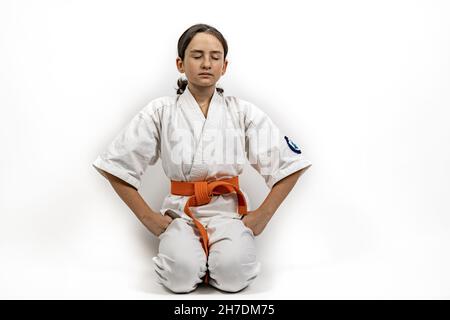 Karate ragazza su sfondo bianco. Una bruna caucasica sportiva in un kimono bianco è impegnata in arti marziali. Medita nella posizione seiza wit Foto Stock