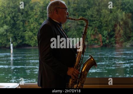 Sassofono suonatore di jazz di fronte a finestre con foresta e fiume blu sullo sfondo. Vista laterale con luce naturale. Foto Stock