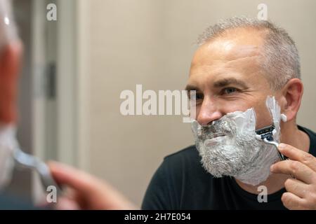 Uomo maturo dai capelli grigi che restyling la sua barba in casa utilizzando il rasoio Foto Stock