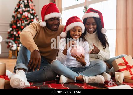 Famiglia nera in babbo natale cappelli mettere moneta in Piggybank Foto Stock