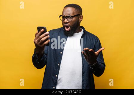 Scioccato stupito uomo afro-americano fissando lo smartphone con ampia bocca aperta isolato su sfondo giallo. Il ragazzo nero sente le emozioni negative, l'espressione facciale stupita Foto Stock