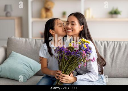 Ti amo, mamma. Bella ragazza asiatica baciare la mamma in guancia e dare mazzo di fiori, seduto sul divano Foto Stock
