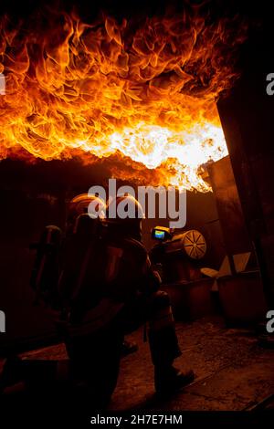 Due vigili del fuoco in una casa piena di fiamme cercando di spegnere il fuoco. Gli uomini indossano dispositivi di protezione respiratoria con caschi, pieni Foto Stock