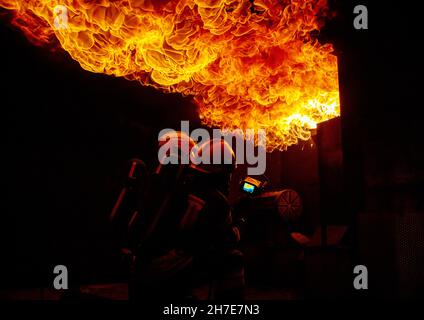 Due vigili del fuoco in una casa piena di fiamme cercando di spegnere il fuoco. Gli uomini indossano dispositivi di protezione respiratoria con caschi, pieni Foto Stock