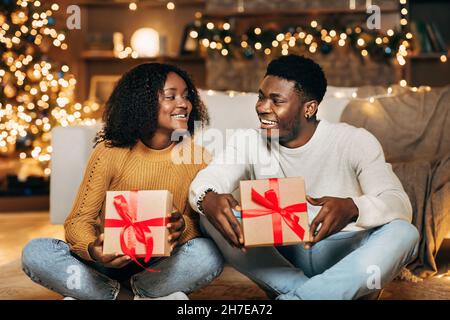 Giovane coppia nera affettuosa che si scambiano regali di Natale in soggiorno decorato Foto Stock