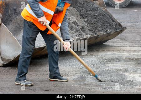 Un operatore su strada in un giubbotto arancione luminoso prepara una vecchia superficie per le riparazioni e rimuove i detriti stradali in una benna livellatrice. Spazio di copia, primo piano. Foto Stock