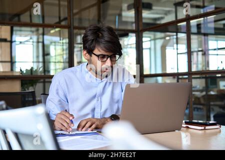 Giovane uomo d'affari indiano che guarda il webinar sul notebook in ufficio. Foto Stock