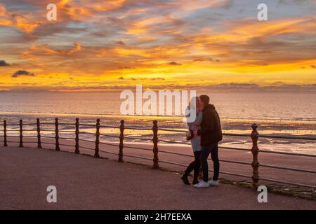 Blackpool, Lancashire. Meteo Regno Unito. 22 Nov 2021. L'amore è nell'aria. Due turisti al resort godono di un super tramonto dopo una fredda giornata luminosa sulla costa. I turisti e i turisti Steve & Gill possono godersi un bacio romantico mentre il sole tramonta sul Mare d'Irlanda. Credit: MediaWorldImages/AlamyLiveNews Foto Stock