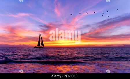 Una barca a vela sta navigando lungo l'oceano con un gregge di uccelli che volano sopra UN cielo colorato di tramonto Foto Stock