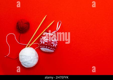 Palla di Natale a maglia e palle di filato con aghi da maglieria su sfondo rosso. Biglietto di Natale, lavoro da ago. Foto orizzontale. Foto Stock