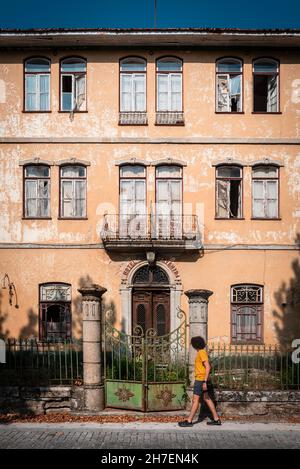 Finestre, porta d'ingresso e recinzione di un antico palazzo abbandonato con un ragazzo che passa sul marciapiede. Foto Stock